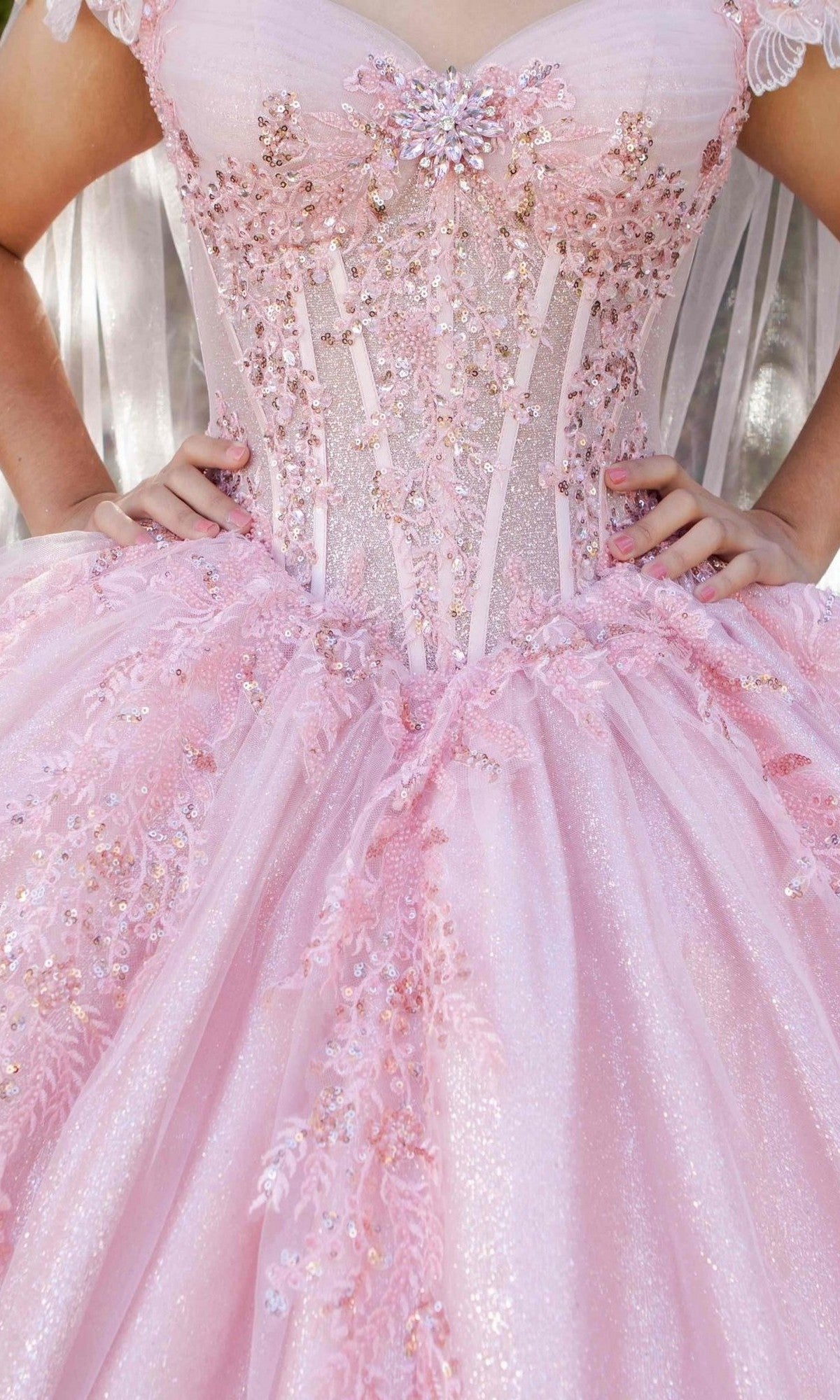 A person wearing the Quinceanera Dress 8270J By Cinderella Couture—a luxurious light pink ball gown embellished with sequins, lace, and 3D floral appliques—stands with hands on hips; upper body visible.